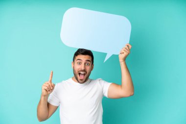 Mavi arka planda izole edilmiş yakışıklı beyaz bir adam elinde şaşırmış bir ifadeyle boş bir konuşma balonu tutuyor.
