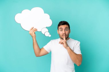 Mavi arka planda izole edilmiş yakışıklı beyaz bir adam düşünme balonu tutuyor ve sessizlik jesti yapıyor.