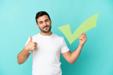 Mavi arka planda izole edilmiş yakışıklı beyaz bir adam elinde başparmağıyla bir kontrol simgesi tutuyor.