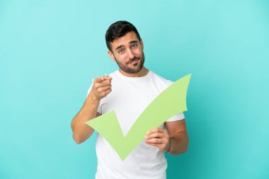 Mavi arka planda izole edilmiş yakışıklı beyaz adam elinde bir çek simgesi tutuyor ve ön tarafı işaret ediyor.