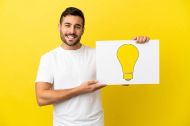 Sarı arka planda izole edilmiş yakışıklı beyaz bir adam elinde mutlu ifadeli bir ampul simgesi olan bir levha tutuyor.