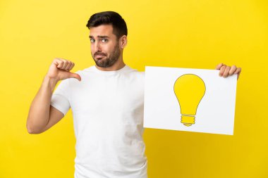 Sarı arka planda izole edilmiş yakışıklı beyaz bir adam elinde gururlu bir jestli ampul simgesi olan bir pankart tutuyor.