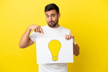 Sarı arka planda izole edilmiş yakışıklı beyaz bir adam elinde ampul simgesi olan bir pankart tutuyor ve onu işaret ediyor.