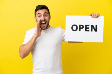 Sarı arka planda izole edilmiş yakışıklı beyaz bir adam. Elinde açık yazılı bir pankart var ve bağırıyor.