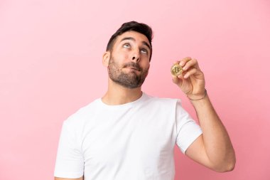 Elinde bir Bitcoin tutan genç adam pembe arka planda izole edilmiş ve yukarı bakıyor