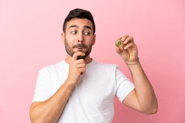 Elinde pembe arka planda izole edilmiş bir Bitcoin tutan genç adamın şüpheleri ve düşünceleri var.