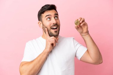 Elinde bir Bitcoin tutan genç adam pembe arka planda izole edilmiş bir fikir ararken bir yandan da