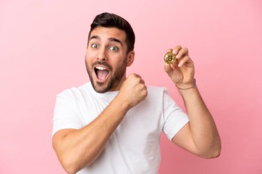 Pembe arka planda izole edilmiş Bitcoin tutan genç adam zaferi kutluyor.