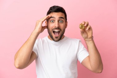 Pembe arka planda, sürpriz bir ifadeyle izole edilmiş bir Bitcoin tutan genç adam.
