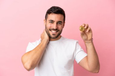 Pembe arka planda izole edilmiş Bitcoin tutan genç adam gülüyor.