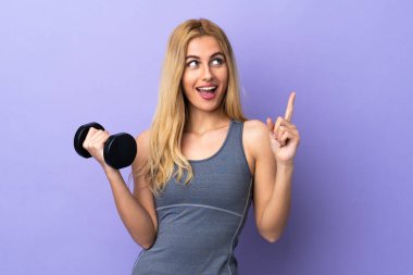 Genç sarışın spor kadın, izole edilmiş mor arka plan üzerinde halter kaldırıyor. Çözümü bulmak için parmağını kaldırıyor.