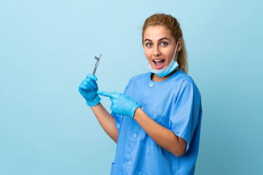 İzole edilmiş mavi arka planda aletleri tutan genç bir dişçi var.