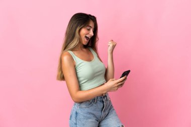 Pembe arka planda izole edilmiş genç beyaz kadın, telefon zafer pozisyonunda.
