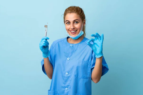 Młoda Kobieta Dentysta Trzyma Narzędzia Odizolowanym Niebieskim Tle Pokazując Znak — Zdjęcie stockowe