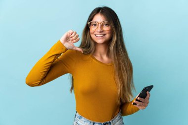 Mavi arka planda izole edilmiş cep telefonu kullanan beyaz bir kadın gururlu ve kendini beğenmiş.