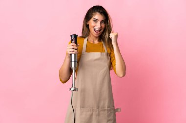 Genç aşçı kadın pembe arka planda izole edilmiş el blenderi kullanıyor. Kazanan pozisyonda zaferi kutluyor.