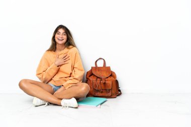 Genç bir kız öğrenci dizüstü bilgisayarla yerde oturuyor. Beyaz arka planda izole edilmiş, gülümsüyor.