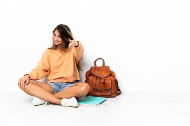 Genç bir kız öğrenci dizüstü bilgisayarla yerde oturuyor beyaz arka planda izole edilmiş, mutlu bir ifadeyle ön tarafı gösteriyor.