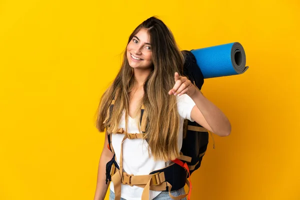大きなバックパックを黄色の背景に幸せな表情で前を指して隔離された若い登山家の女性 — ストック写真