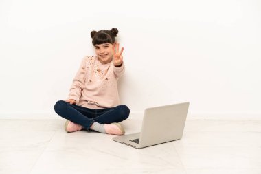 Dizüstü bilgisayarı olan küçük bir kız yerde oturmuş mutlu ve parmaklarıyla üçe kadar sayıyor.