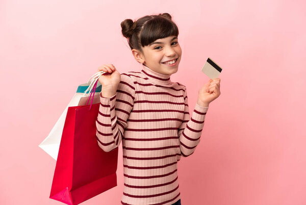 Маленькая белая девочка, изолированная на розовом фоне, держащая сумки с покупками и кредитку