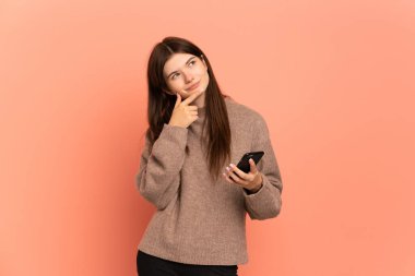 Genç Ukraynalı kız pembe arka planda izole edilmiş cep telefonu kullanıyor ve düşünüyor.