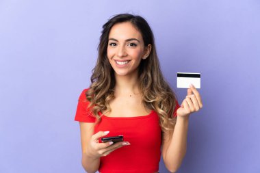 Mor arka planda izole edilmiş genç beyaz bir kadın kredi kartıyla cep telefonuyla alışveriş yapıyor.