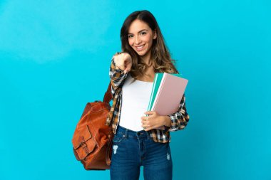 Genç bayan öğrenci mavi arka planda izole edilmiş, mutlu bir ifadeyle ön tarafı işaret ediyor.
