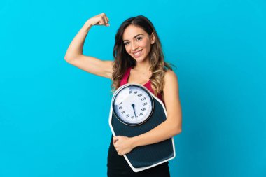 Elinde bir tartı tutan beyaz bir kadın mavi arka planda izole edilmiş bir tartı tutuyor ve güçlü bir hareket yapıyor.