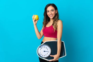 Mavi arka planda tartı tutan beyaz bir kadın. Tartı makinesiyle ve elmayla izole edilmiş.