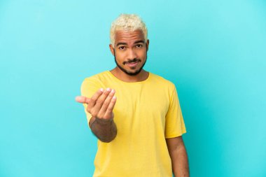 Genç Kolombiyalı yakışıklı adam, mavi arka planda izole edilmiş, bizimle gelmesi için davet ediliyor. Gelmene sevindim.