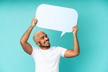 Genç Kolombiyalı yakışıklı adam mavi arka planda izole edilmiş boş bir konuşma balonu tutuyor.