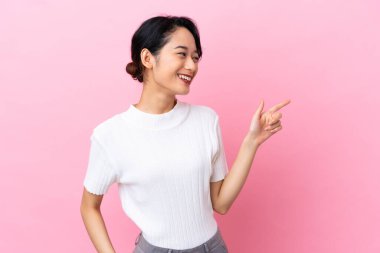 Pembe arka planda izole edilmiş genç Vietnamlı bir kadın parmak göstererek bir ürün sunuyor.