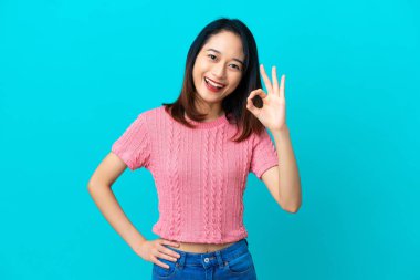 Mavi arka planda izole edilmiş genç Vietnamlı kadın parmaklarıyla tamam işareti gösteriyor.