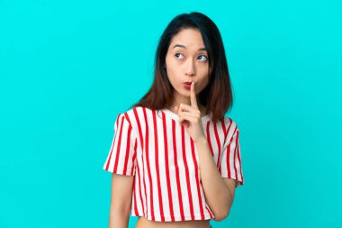Mavi arka planda izole edilmiş genç Vietnamlı kadın sessiz bir jest işareti gösteriyor. Parmağını ağzına sokuyor.