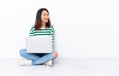 Yerde dizüstü bilgisayarı olan genç Vietnamlı bir kadın izole edilmiş beyaz duvarda mutlu ve gülümsüyor.