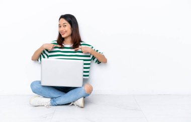Yerde dizüstü bilgisayarı olan genç Vietnamlı bir kadın kendini beğenmiş ve gururlu bir şekilde beyaz duvarda tek başına oturuyor.