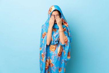 Geleneksel kostümlü genç Faslı kadın yorgun ve hasta bir ifadeyle mavi arka planda izole edilmiş.