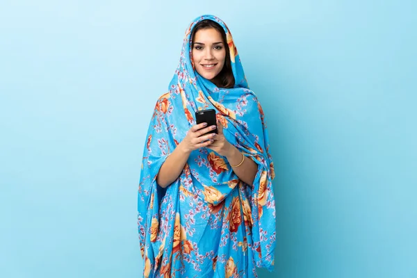 年轻的摩洛哥女人穿着传统服装 背景是蓝色的 她用手机传达了一个信息 — 图库照片