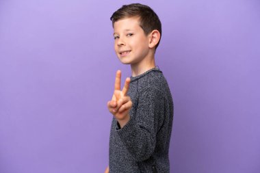 Mor arka planda izole edilmiş küçük çocuk gülümsüyor ve zafer işareti gösteriyor.