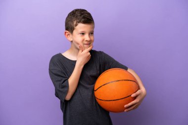 Mor arka planda izole edilmiş basketbol oynayan küçük çocuk yana bakıyor ve gülümsüyor.