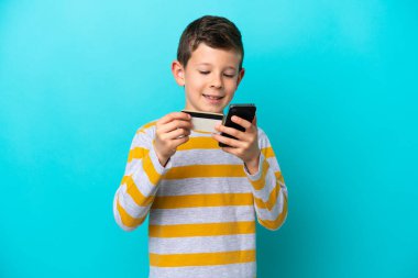 Küçük çocuk mavi arka planda izole edilmiş, kredi kartıyla cep telefonuyla alışveriş yapıyor.