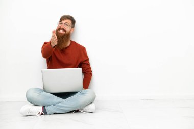 Dizüstü bilgisayarı olan genç, beyaz arka planda izole edilmiş beyaz bir adam iyi bir anlaşma için el sıkışıyor.