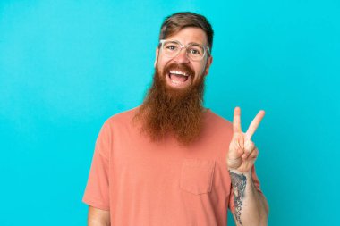 Kırmızımsı beyaz adam mavi arka planda izole edilmiş gözlüklü ve tamam işareti yapıyor.