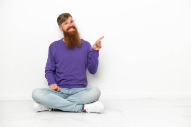 Beyaz arka planda izole bir şekilde yerde oturan beyaz tenli, kırmızımsı genç bir adam var.