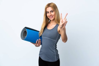 Genç sporcu kadın yoga derslerine giderken izole edilmiş beyaz arka planda bir paspas tutuyor gülümsüyor ve zafer işareti gösteriyor.