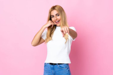 Genç Uruguaylı sarışın kadın soyutlanmış pembe arka planda telefon jesti yapıyor ve ön tarafı işaret ediyor.