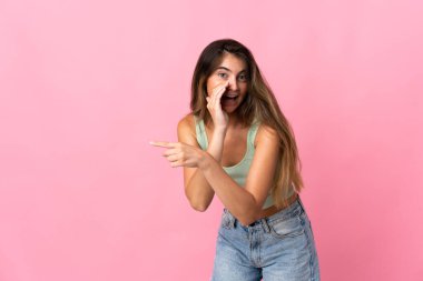 Pembe arka planda izole edilmiş genç beyaz kadın bir ürünü sunmak ve bir şeyler fısıldamak için kenarı işaret ediyor.