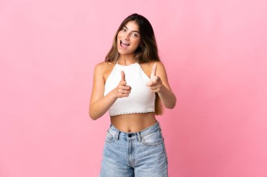 Pembe arka planda izole edilmiş genç beyaz kadın ön tarafı işaret ediyor ve gülümsüyor.