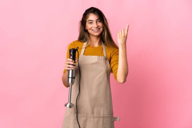 Genç aşçı kadın pembe arka planda izole edilmiş el blenderi kullanıyor. Harika bir fikre işaret ediyor.